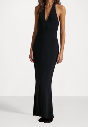 Femme portant une longue robe noire dos-nu avec un décolleté en V profond, debout devant un fond blanc uni.