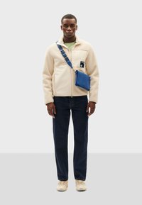 Cremfarbene Fleecejacke mit Reißverschluss und Tasche, kombiniert mit dunkelblauen Jeans und beigefarbenen Sneakers, accessoriert mit einer blauen Umhängetasche.