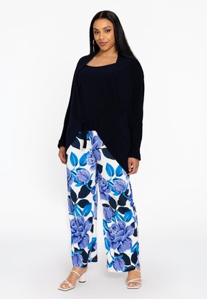 Ein marineblauer Cardigan über einem dunklen Oberteil, kombiniert mit weit geschnittenen Hosen, die ein großes blaues Blumendesign auf weißem Grund zeigen.