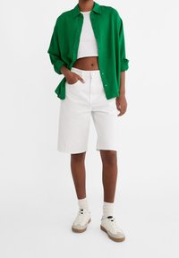 Groene button-up shirt over een witte crop top, gecombineerd met knie-lange witte shorts en beige sneakers met crèmekleurige sokken.