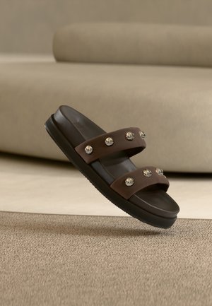 Καφέ σανδάλι slide με δύο λουράκια που διαθέτουν στρογγυλά ασημένια καρφίτσια, εμφανίζεται σε μπεζ χαλί με μπεζ καναπέ στο φόντο.