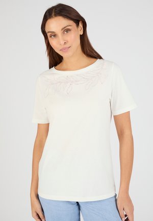 Femme aux longs cheveux foncés portant un haut blanc à manches courtes avec une broderie subtile de feuilles près de l'encolure et un jean bleu clair.