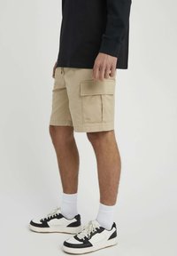 Shorts cargo beiges avec plusieurs poches, fabriqués en coton, avec une coupe décontractée. Associés à des baskets noires et blanches et des chaussettes blanches.