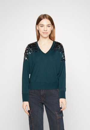 Pull en teal avec un col en V et des embellissements de sequins noirs sur les épaules. Manches longues et coupe décontractée, accompagné de jeans foncés.