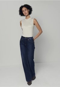 Haut sans manches beige clair, associé à un jean large bleu foncé. La tenue comprend des détails de couture subtils et est portée avec des bottes marron.