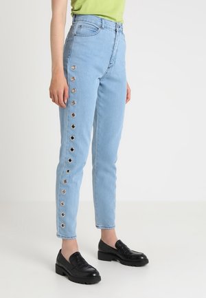 Persoon draagt lichtblauwe high-waist jeans met metalen oogjes langs de buitenste beennaden, gecombineerd met zwarte loafers en een groene top.