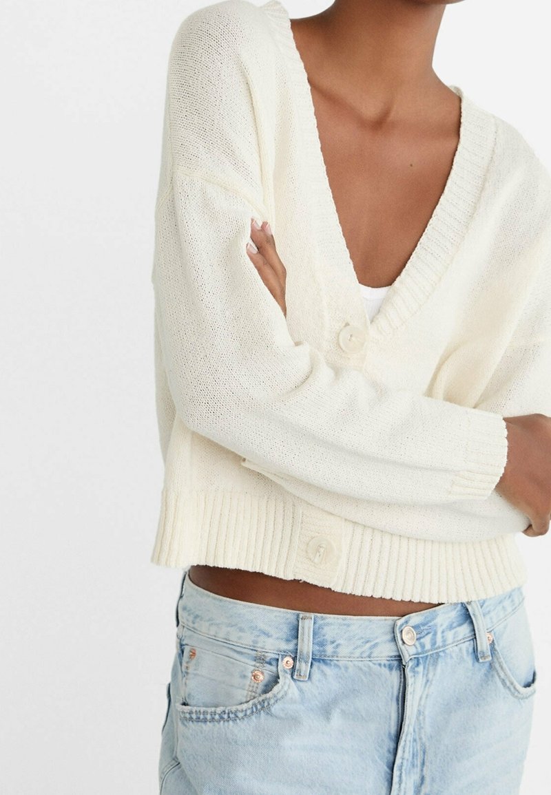 Light cream cardigan with a deep V-neck, textured knit fabric, ribbed cuffs, and two buttons at the front, paired with light blue jeans.