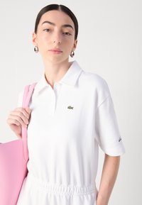 Balta polo marškinėliai su trumpomis rankovėmis ir apykakle, su žalia siuvinėta krokodilo logotipu. Derinama su rožine rankine ir sidabriniais auskarais žiedais.