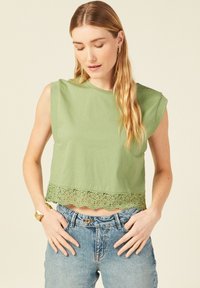 Top verde corto realizzato in tessuto morbido, con orlo in pizzo a scallop. Il design include maniche corte arrotolate e una vestibilità rilassata.