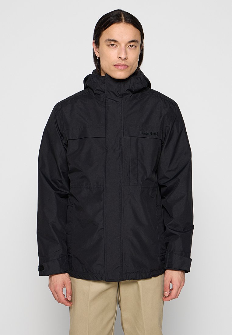 Chaqueta impermeable negra con cremallera en la parte delantera, dos bolsillos en el pecho, capucha ajustable y puños elásticos para un ajuste ceñido. Textura suave.