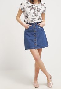 Žena v bílé blůze s potiskem motýlů, zastrčené do denimu sukně s knoflíky, stojící s překříženýma nohama a rukama v kapsách.