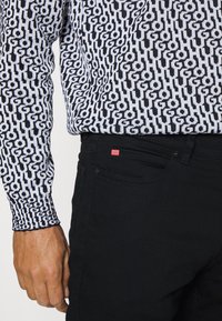 Primo piano di una persona che indossa una camicia a maniche lunghe a righe bianche e nere infilata in pantaloni neri con un piccolo etichetta rossa sulla tasca.