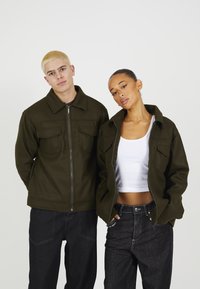 Olivenfargede jakker i teksturert stoff, med glidelås og frontlommer, iført sammen med svarte denimbukser og en hvit topp.