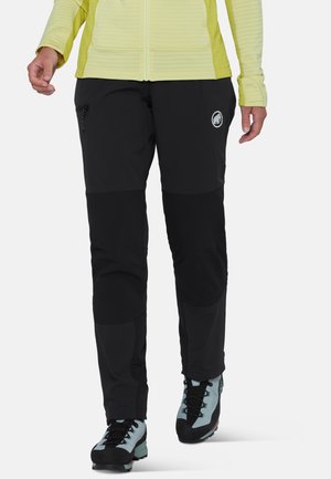 Persona che indossa pantaloni outdoor neri con un logo, scarponi da trekking azzurri e una giacca gialla con zip, camminando verso avanti su sfondo bianco.