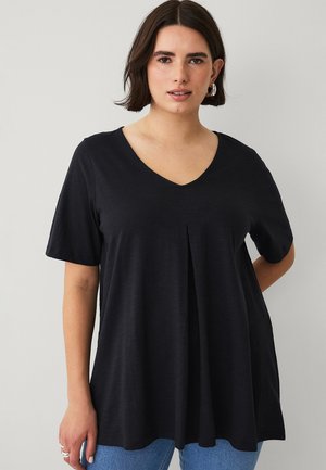 Vrouw die een zwarte V-hals top met korte mouwen en een plooitje middenvoor draagt, gecombineerd met een blauwe spijkerbroek, staand tegen een effen lichte achtergrond.