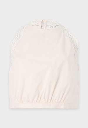 Femeie care poartă un top crem fără mâneci și pantaloni largi bej, ținând o geantă maro închis cu mâner superior.