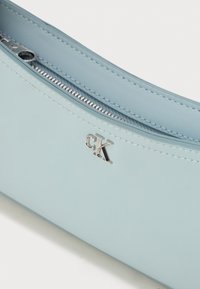 Lysblå lædertaske med en glat overflade, præget af en sølvfarvet lynlås og CK-logo. Enkel, slank design med buede kanter.