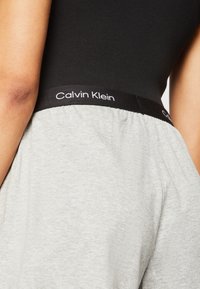 Camiseta sin mangas gris con una banda elástica negra que presenta el logo de "Calvin Klein". La tela parece suave con una textura acanalada.