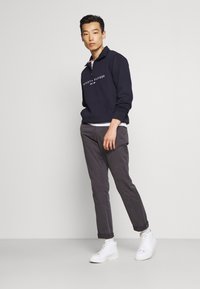 Sudadera de forro polar azul marino con cremallera de un cuarto y logo blanco, combinada con pantalones gris ajustados y zapatillas blancas sobre un fondo neutral.
