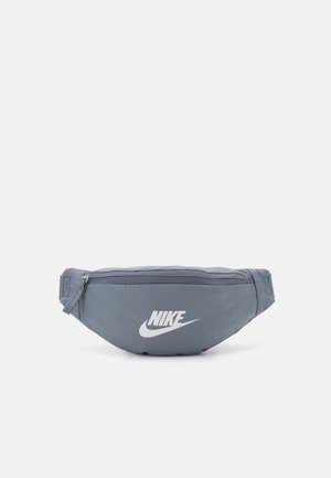 Riñonera gris con bolsillo con cremallera y logotipo blanco de Nike centrado en el frontal sobre un fondo liso.