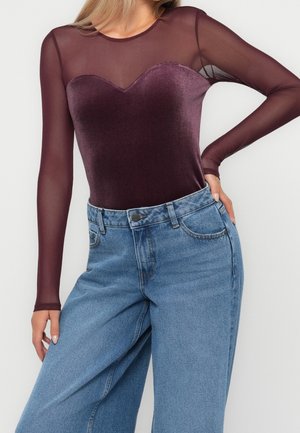 Femme portant un body ajusté en velours violet avec des manches longues transparentes et un jean bleu ample taille haute.