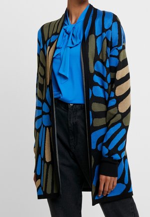 Person iført en åben cardigan med sort, blå, grøn og beige mønster over en lyseblå bluse med sløjfe, kombineret med sorte bukser.