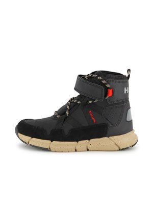 Zwarte atletische mid-top schoen met een gestructureerde bovenkant, verstelbare strap, reflecterende accenten en een lichtbruine rubberen zool met spikkels.