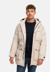 Giacca invernale beige imbottita con cappuccio, dotata di quattro tasche frontali, chiusura con zip e una texture morbida. Indossata sopra un maglione scuro.
