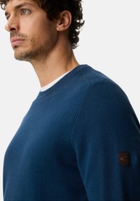 Men’s blue textured sweatshirt with a crew neck, featuring a contrasting white inner collar and a brown logo patch on the left sleeve.