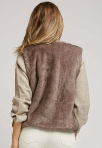 Femme aux longs cheveux blonds portant un pull beige à manches retroussées et un gilet en peluche brun, de dos contre un fond uni.