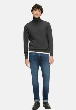Maglione grigio a collo alto con fondo a costine, indossato sopra una camicia bianca. Abbinato a jeans blu aderenti e stivaletti neri in pelle.