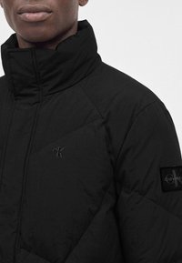 Veste matelassée noire à col montant portée par une personne, avec petits logos Calvin Klein brodés et en patch sur la poitrine et la manche.