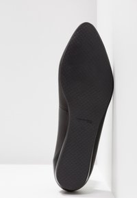 Semelle de chaussure noire à bout pointu avec motif texturé en losanges, appuyée contre un coin entre un mur blanc et un sol blanc.