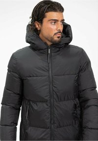 Veste en duvet noire avec un col montant et une capuche attachée. Caractéristiques : fermeture éclair à l'avant, poches latérales et sections matelassées pour l'isolation.