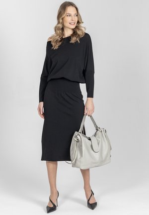 Kobieta w czarnej bluzce z odkrytymi ramionami i spódnicy, trzymająca dużą jasnoszarą torebkę, ubrana w czarne szpilki, stoi na jasnym tle.