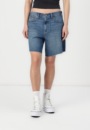 Shorts en denim bleu moyen avec un ourlet effiloché. Présente un design classique à cinq poches et une fermeture par bouton à l'avant. Assortis avec des baskets blanches.