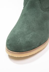 Bottines en suède vertes avec un bout rond, dotées d'une semelle en caoutchouc et de coutures minimales. Texture lisse et éclat subtil.