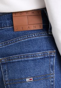 Jeans de mezclilla con una etiqueta de cuero marrón grabada con "TOMMY JEANS". Tela azul con costuras amarillas y un pequeño logo tricolor en el bolsillo trasero.