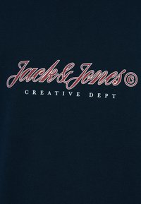Μπλε μπλουζάκι ναυτικού με το λογότυπο "Jack & Jones" σε κόκκινη γραφική γραφή και τη φράση "CREATIVE DEPT" σε λευκά γράμματα από κάτω, κατασκευασμένο από μαλακό βαμβακερό ύφασμα.