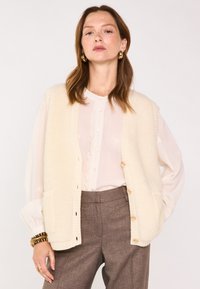 Gilet de couleur crème avec une texture douce et duveteuse, doté d'une bordure en tricot côtelé, de boutons à l'avant et de poches. Porté par-dessus une blouse légère et un pantalon.