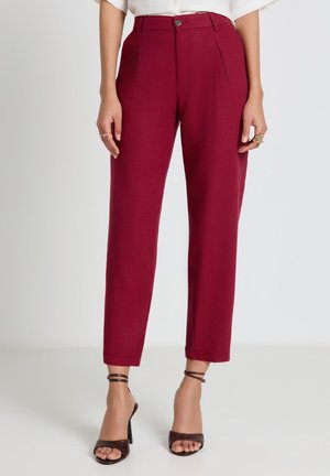 Femme portant un pantalon bordeaux taille haute et fuselé, et des sandales à talons hauts noires avec une bride à la cheville, debout devant un fond uni.