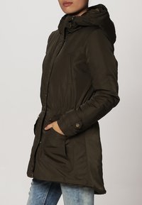 Parka matelassée vert olive avec capuche, fermeture éclair et boutons à l'avant, taille élastique, et deux poches avant avec rabats. Texture lisse.