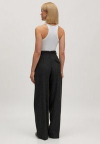 Vrouw met kort blond haar, gekleed in een mouwloze witte top en hoog-modische zwarte wijde broek, staand tegen een effen achtergrond.