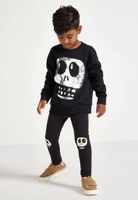 Young boy wearing black sweatshirt with large silver skull and black pants with small skulls on knees, standing on white floor.