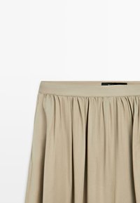 Jupe plissée beige avec une large ceinture, tissu froncé créant de doux plis, sur un fond blanc uni.