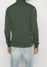 Dlouhý rukáv, tmavě zelená polo košile z měkké látky, s límečkem, bočními rozparky a přiléhavým střihem. Nosit s světle modrými džínami.