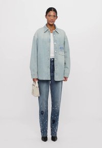 Gai zila džinsu oversized krekls, balta bluze, zili džinsi ar pērļu rotājumiem un melnas norādītas kurpes. Mazs balts rokassoma ar akcentu.