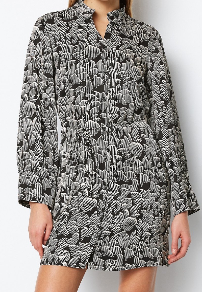 Schwarzes Kleid mit einem Blättermuster in Weiß und Grau. Verfügt über einen Kragen, lange Ärmel und eine taillierte Form mit einem gebundenen Gürtel. Seidene Textur.
