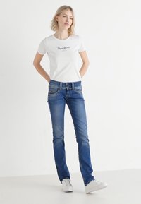 Hvit t-skjorte med kort erme og logo, sammen med blå denimjeans. Modellen har på seg hvite joggesko. Ensfarget bakgrunn.