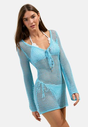 Noor naine, kes kannab hele sinist avara kudumiga pikka varrukaga kleiti valge lainelise servaga bikiini peal, seisab ühevärvilise heleda tausta ees.